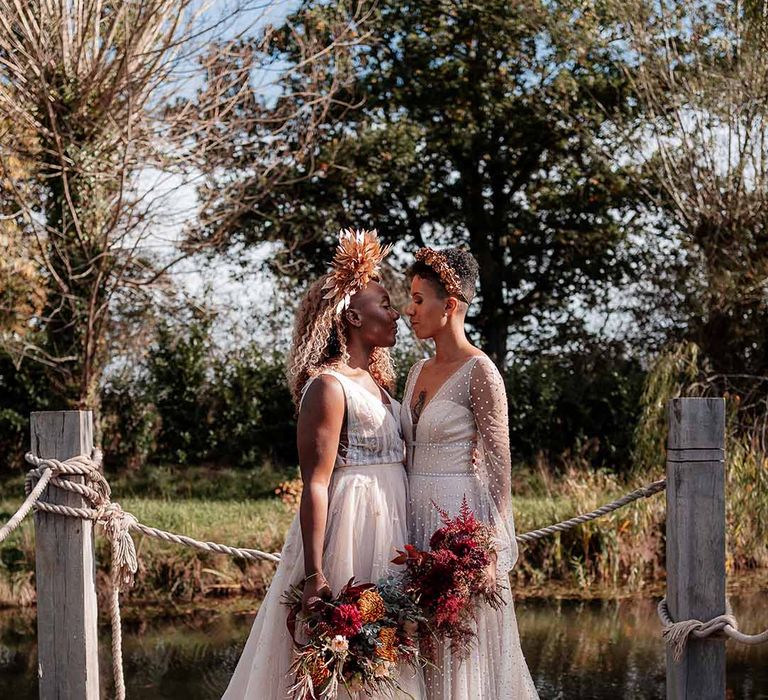 Bride in crystal embellished gown and gold headdress embracing bride in sleeveless floaty peach gown holding dried autumnal floral bouquets on a dock by the river at Silchester Farm