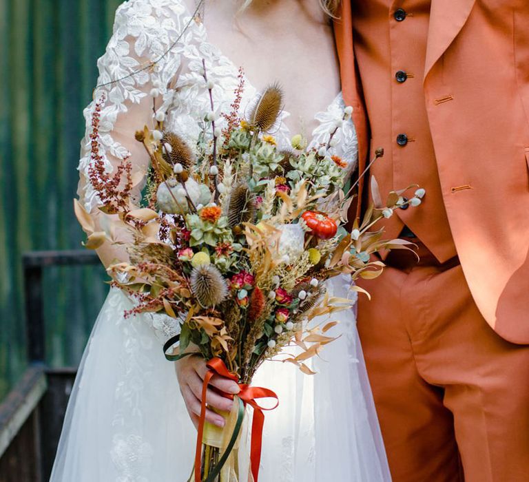 Bride holding dried flower autumnal bouquet with bouquet with white dried cotton stems, terra cotta celosia seeds, green foliage wearing a long sleeved v neck soft ivory dress with lace floral detailing on the bodice and sheer sleeves and headpiece with delicate details and mix of soft gold & bronze metals and groom in burnt orange rust coloured suit with brown tie, pocket square, brown accessories and dried flower, rose and pampas grass boutonniere  