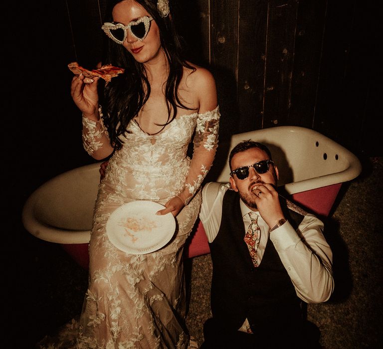 Bride in off the shoulder Abigail Westrup bridal gown with long, sheer sleeve and lace detailing wearing cat eye bejewelled sunglasses sitting on the edge of a bathtub with groom in black waistcoat and flower patterned tie wearing Raybans sunglasses both eating pizza 