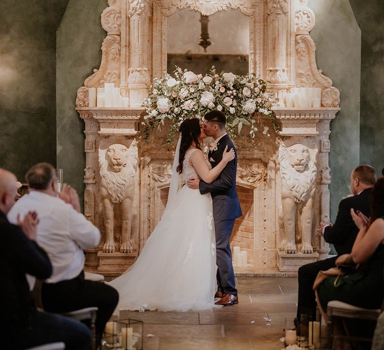 Bride & groom kiss in front of pale pink and green floral installation above fireplace 