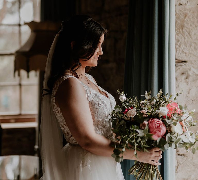 Bride wears her brown hair in loose curls and holds pink and white floral bouquet 