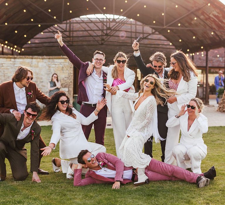 Bridal party striking a pose: Bridesmaids in bridesmaid jumpsuits with white blazers, groomspeople in mismatched groom suits, bride in long sleeve boho lace wedding dress, all in sunglasses, smiling and cheering 