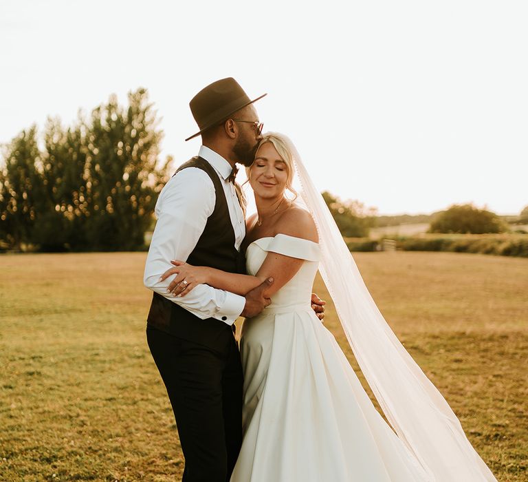 Groom in fedora and black waistcoat with white shirt kisses his bride in off-the-shoulder princess wedding dress