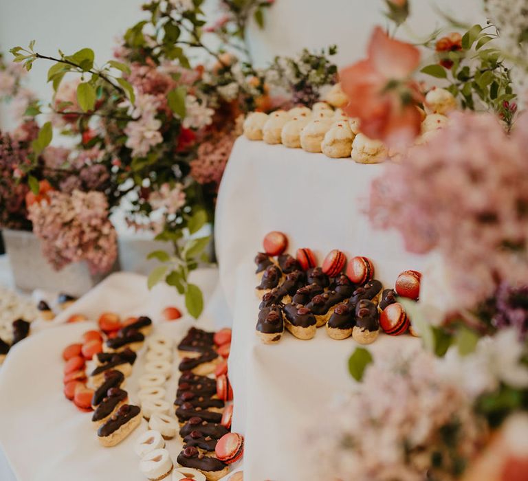 Dessert table complete with mini eclairs and colourful macaroons surrounded by bright florals 