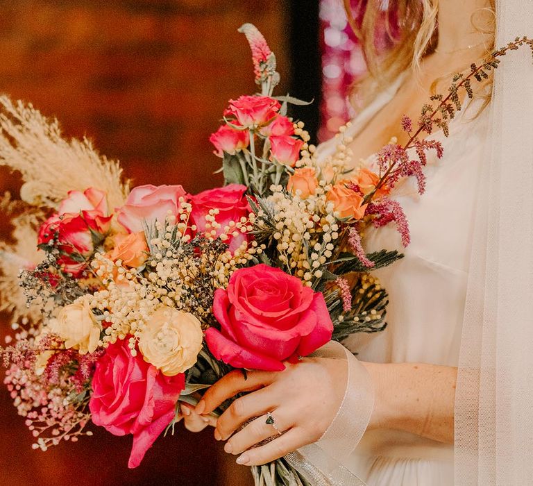 Bride in three quarter length sheer sleeve wedding dress with button and cinched waist details holding pink garden rose, pampas grass, carnations, baby's-breath, dried flower and foliage bridal bouquet with sheer white lace ribbon