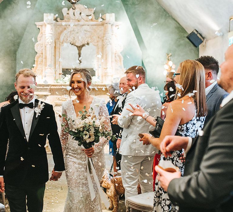 Groom in black tie walks back down the aisle with the bride in a long sleeve 3D lace wedding dress for their confetti moment 