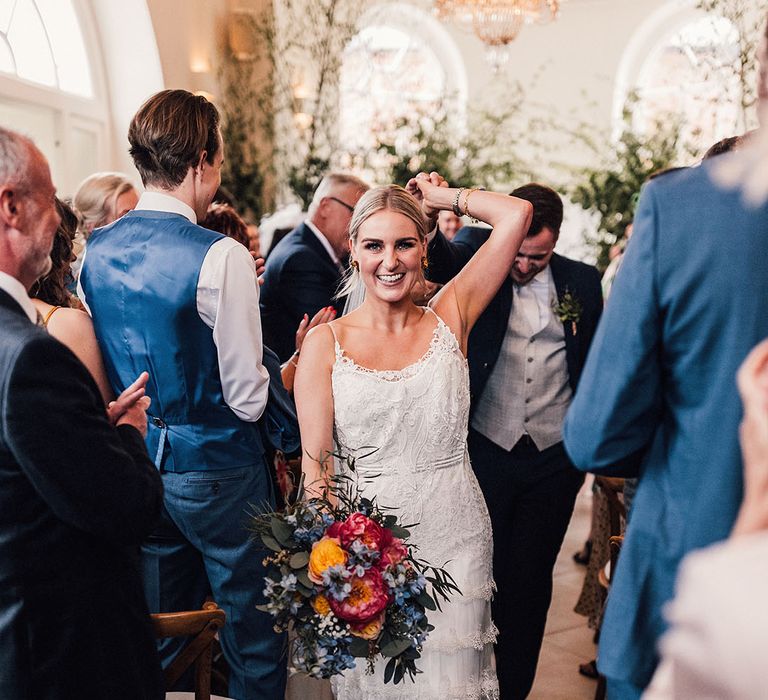 The bride in a lace overlay wedding dress holding a pink and blue wedding bouquet walks back down the aisle with the groom