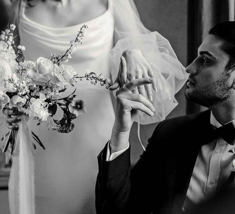 Groom in classic black tuxedo, boutonniere and black bowtie posing with bride in long sheer puff sleeve satin wedding dress with cowl neck