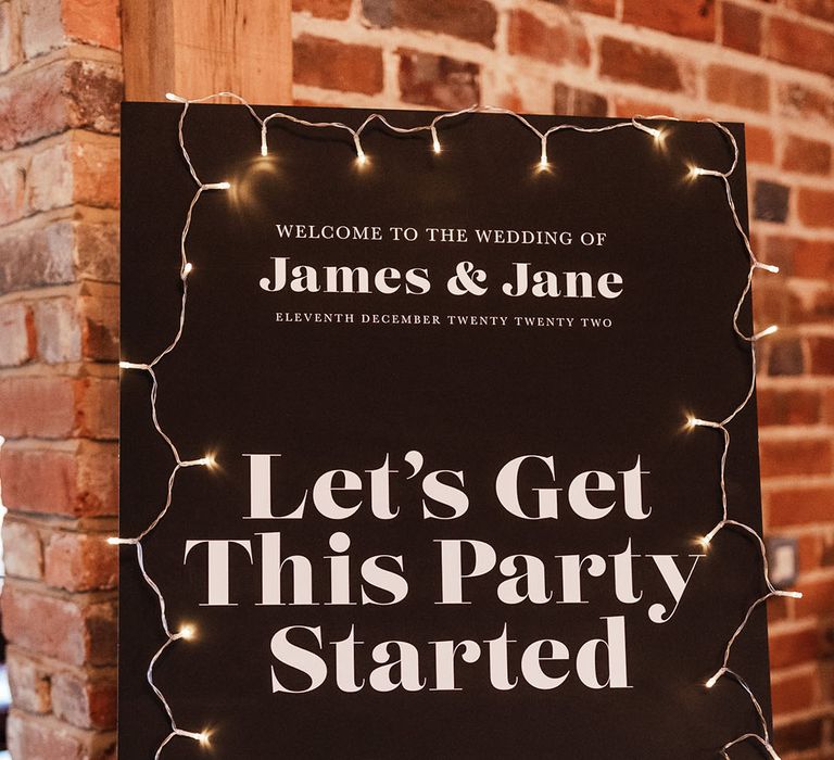 Black and white modern wedding welcome sign with fairy lights decorating the signage on a wooden easel 