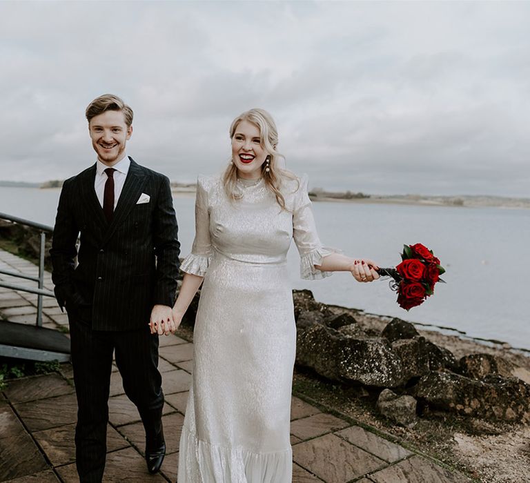 Groom in dark suit with red tie walking with bride in sparkly silver wedding dress holding red rose bouquet 