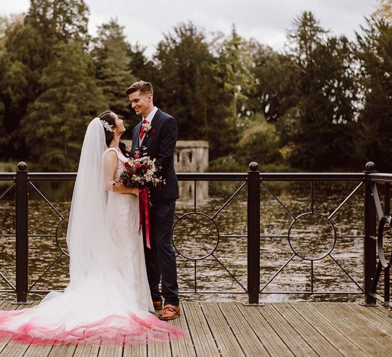 Bride in sleeveless lace wedding dress and church length red ombre veil holding white and red garden rose, peony, eucalyptus and dried foliage flower arrangements with red lace tie standing with groom in deep blue grooms blazer, grey waistcoat, red tie, red pocket square and mixed dried flower boutonniere overlooking the lake