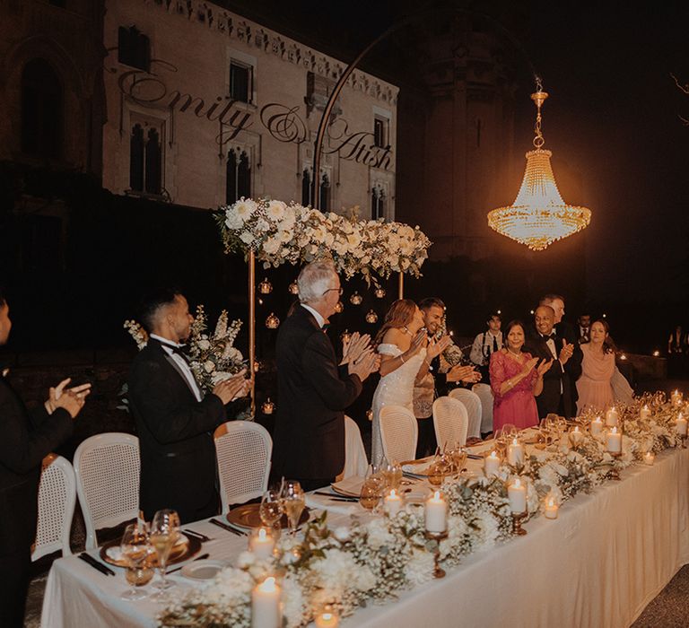 Guests standing by outdoor wedding classic tablescape - white tablecloth, bejewelled outdoor chandeliers, peony and foliage table runners, pillar candles and gold cutlery and crockery 