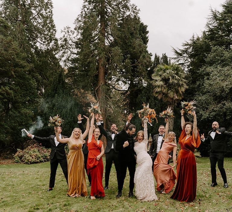 Groomsmen in black tie with the bridesmaids in mismatched bridesmaid dresses in autumnal shades posing with smoke bombs 