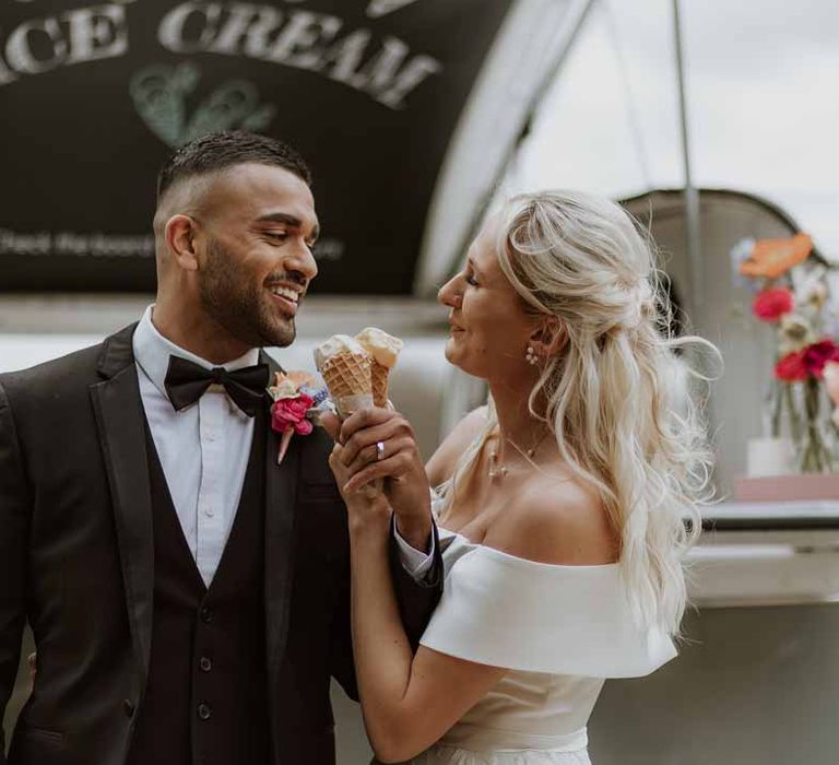 Bride in off the shoulder lace wedding dress eating wedding ice cream with groom in classic three piece grooms tuxedo with colourful spring boutonniere 