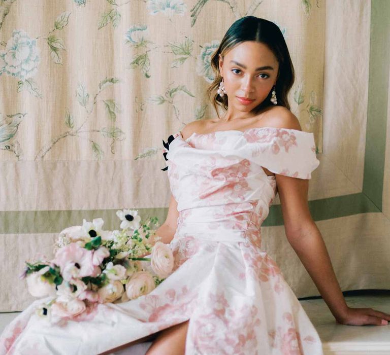 Bride in off the shoulder white and pink floral patterned bridal gown with black bow and front slit holding bridal bouquet with white poppies, light pink Persian buttercups, baby's-breath and foliage at Settrington Orangery 