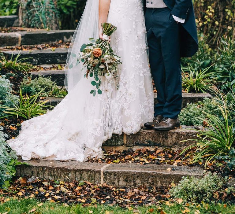 Groom in navy grooms morning suit with grey waistcoat, off-white tie and mixed wildflower boutonniere standing with bride in a-line tulle wedding dress with beaded lace applique with petals and a square neckline with side tulle cutouts holding pampas grass, eucalyptus, blush garden roses, dried flowers and foliage bridal bouquet