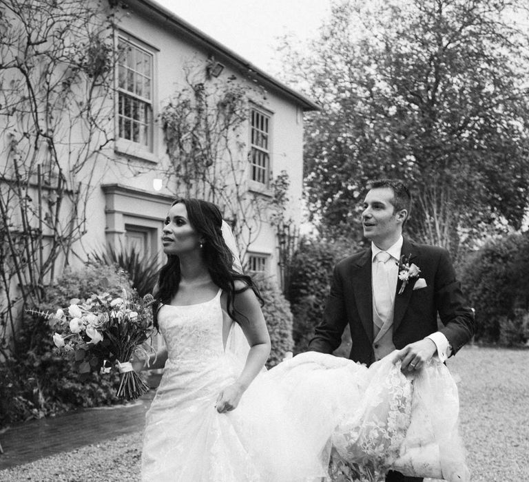 Groom in navy grooms morning suit with grey waistcoat, off-white tie and mixed wildflower boutonniere helping bride with train and veil at South Farm wedding venue