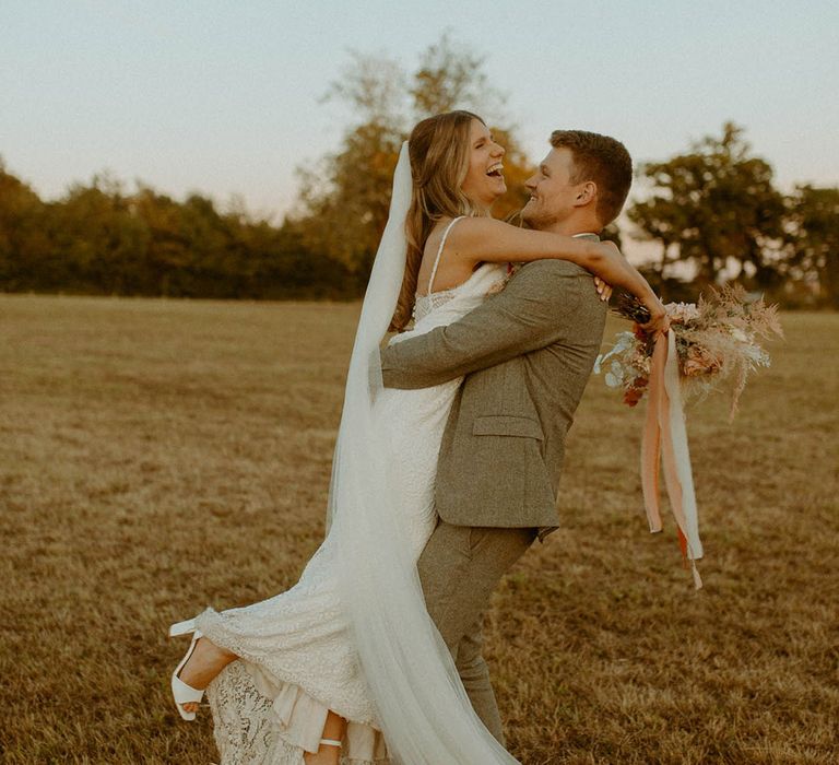 Bride in fitted lace wedding dress being lifted by the groom in grey suit carrying dried wedding flower bouquet for boho wedding 