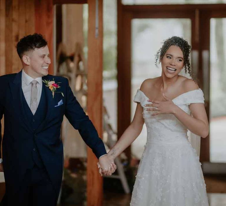 Groom in navy groom morning suit with grey ties and blush rose boutonniere walking into the reception room of Elmore Court with bride in Suzanne Neville off shoulder 3D floral applique wedding dress