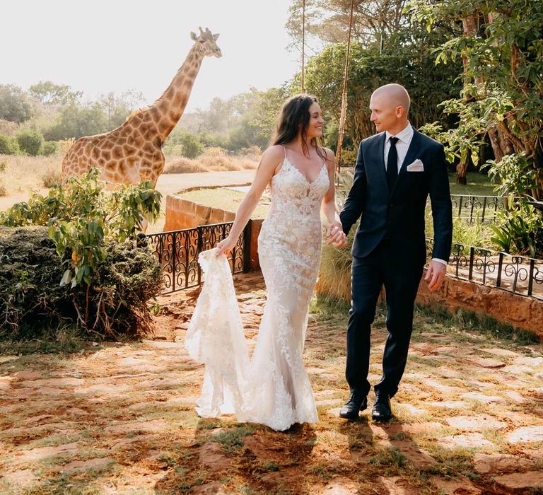 Groom in classic black grooms suit with white pocket square walking hand in hand with lace sleeveless wedding dress with puddle train on the grounds of Giraffe Manor wedding venue with giraffes in the background 