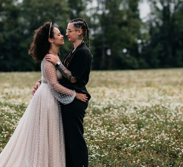 Bride in silver chains, glasses, black shirt and black trousers embracing bride in long sleeve v-neck sheer polka dot overlay sparkly wedding dress and gold celestial bridal headband at at-home wedding ceremony 