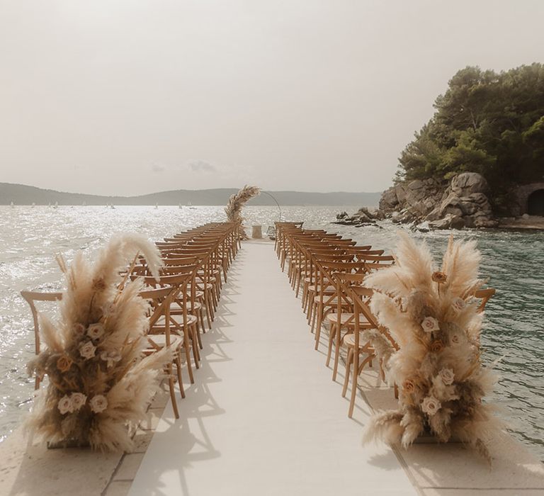 coastal wedding ceremony by the sea with long aisle with wooden chairs, pampas grass floral arrangements and moon gate altar