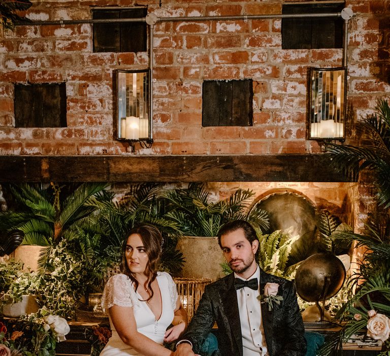 Bride and groom seated wedding photography bride wears jimmy choo gold wedding shoes and silk wedding dress, groom wears - Dolce & Gabanna suit. Exotic green background, rustic side table and cosy candles