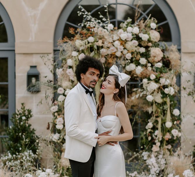 Stylish bride in a satin wedding dress with cup detail and thin straps wearing red lipstick and a bow headdress looking at her groom in a white tuxedo jacket 