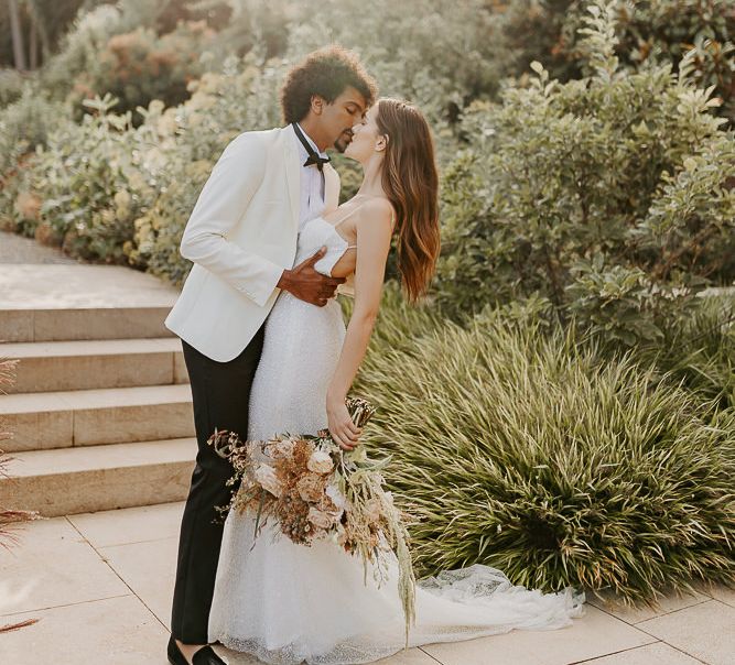 Black groom in a tuxedo with white jacket kissing his bride in a white sequin wedding dress with thin straps 