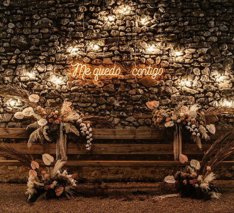 Wooden bench decorated in boho wedding flowers in natural and blush tones, plus, a hanging neon sign and festoon lights 