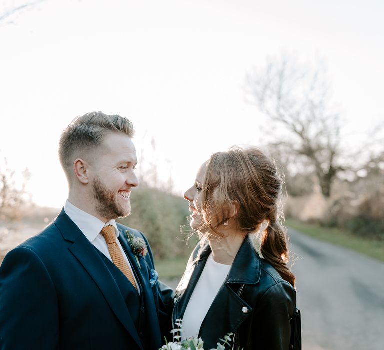 White blonde bearded groom in a navy suit and orange tie smiling at his white red headed bride in a leather jacket 