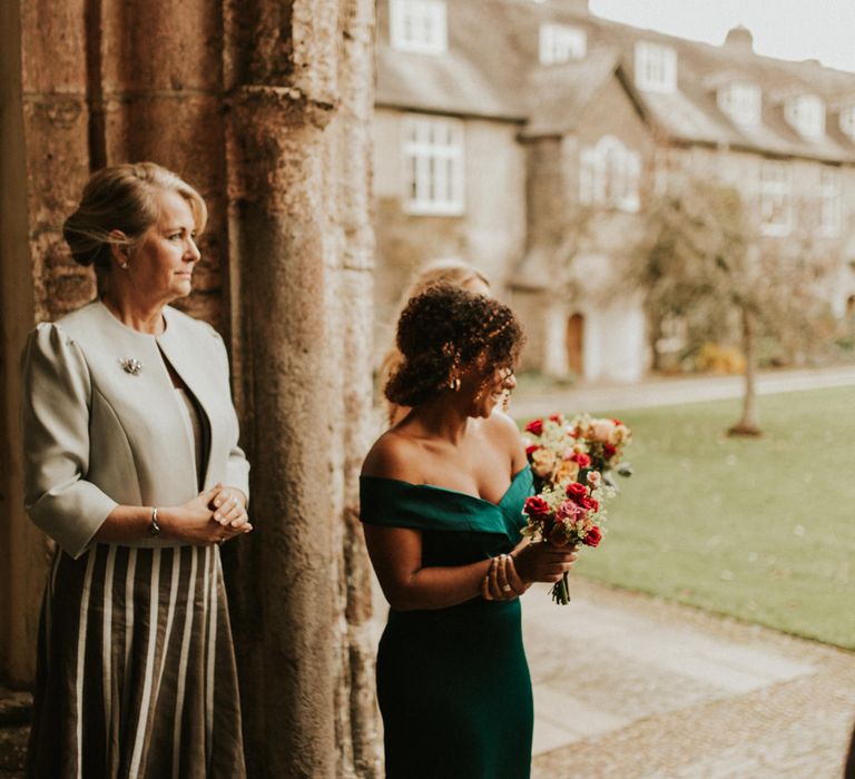 Black bridesmaid with curly hair in a dark green off the shoulder dress holding a red flower bouquet 