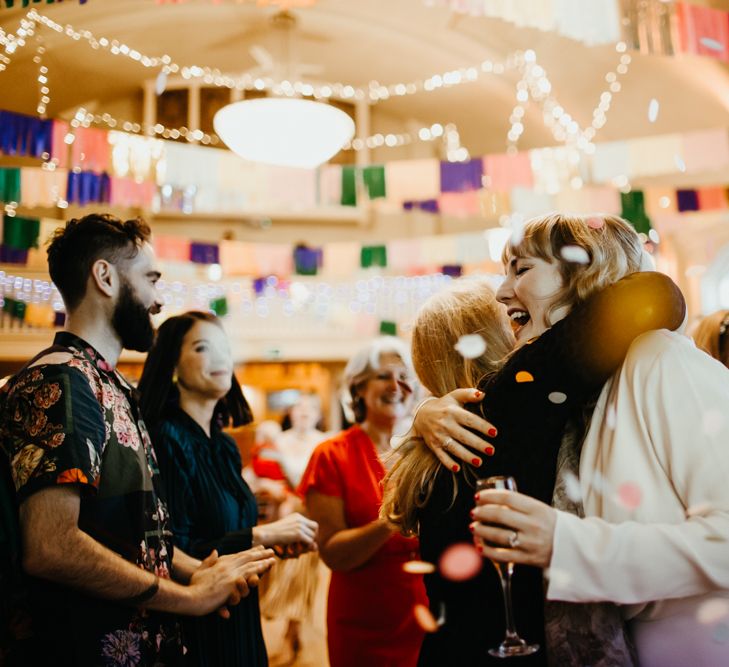 Bride in a long sleeve jumpsuit with red nail polish embracing a wedding guest during the confetti moment 