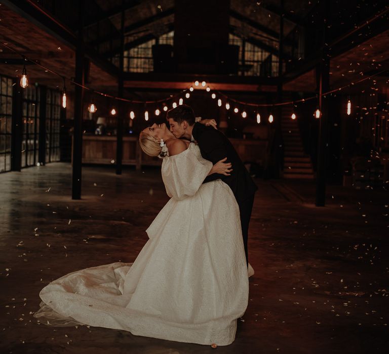 Groom kissing his brides neck in a full skirt wedding dress with blouson sleeves under a canopy of festoon lights 