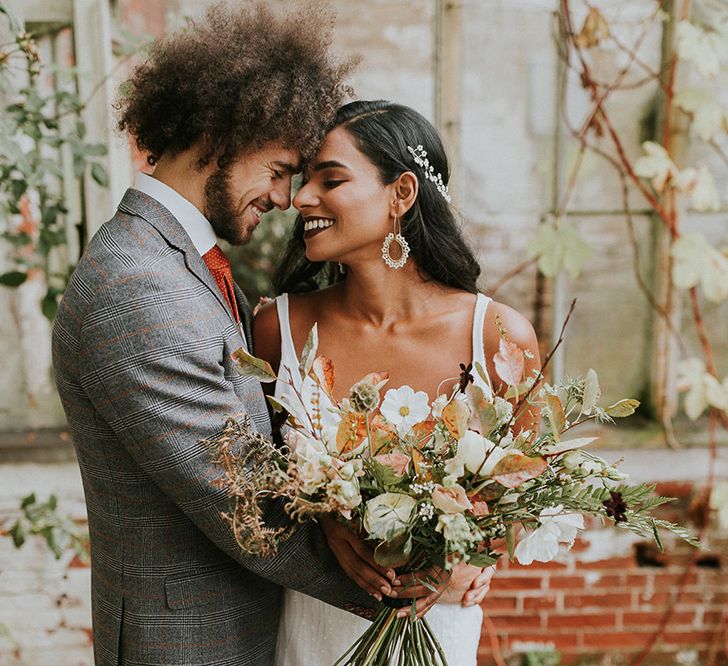 A couple embrace for wedding photos. He wears a grey suit and she holds a bouquet.