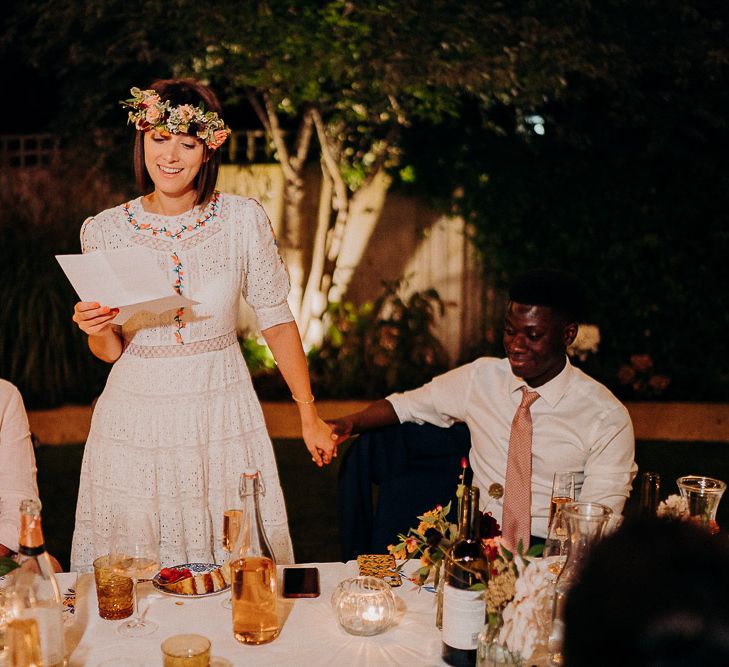 Bride in a white Broderie Anglaise dress with colourful embroidery ready her wedding speech at an outdoor reception 