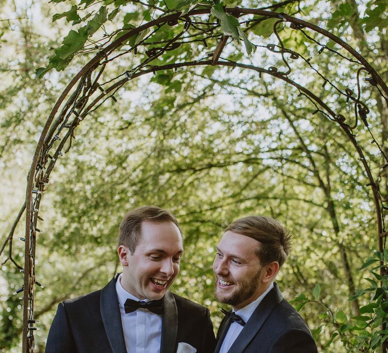 Grooms stand under green floral archway on wedding day