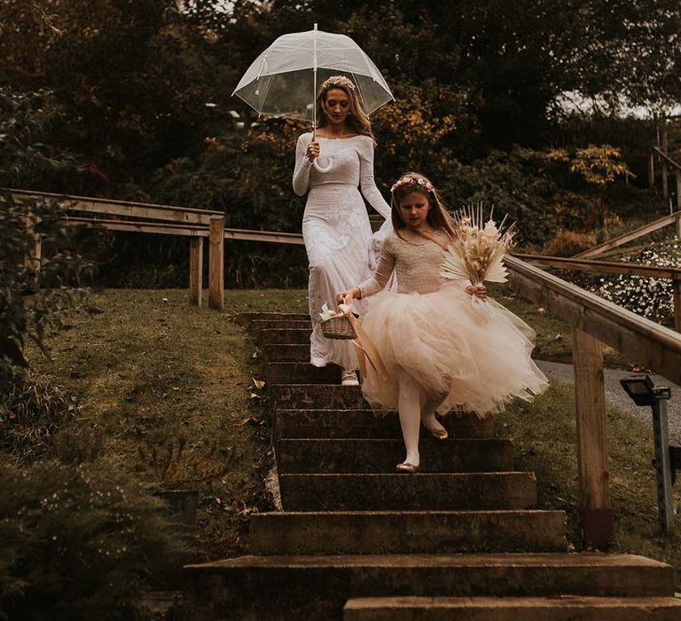 Bride in a fitted wedding dress holding a clear umbrella walking down the steps with her daughter in a peach tulle tutu skirt