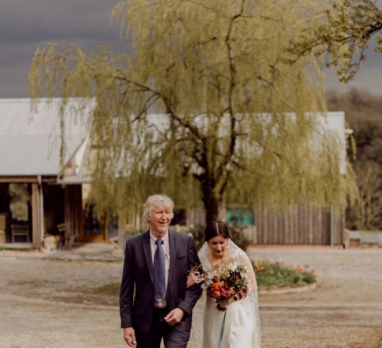 Bride in Charlie Brear wedding dress and chapel length daisy applique veil holding multicoloured bridal bouquet walks arm-in-arm with male wedding guest