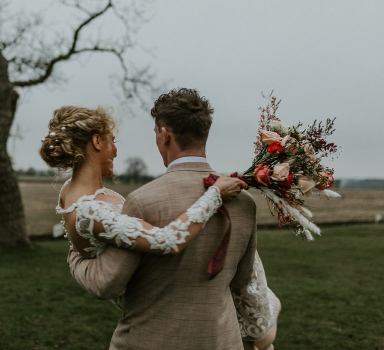 Groom carrying the bride through a field, she is wearing a long sleeved gown with floral embroidered pattern