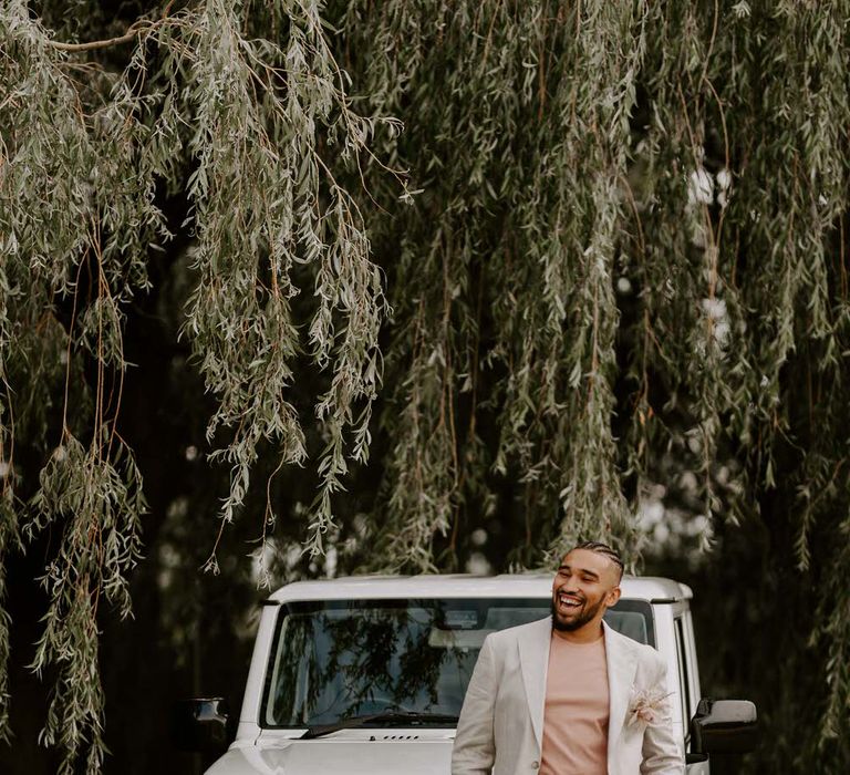 Groom in a cream suit leaning on a large white 4x4 Jeep