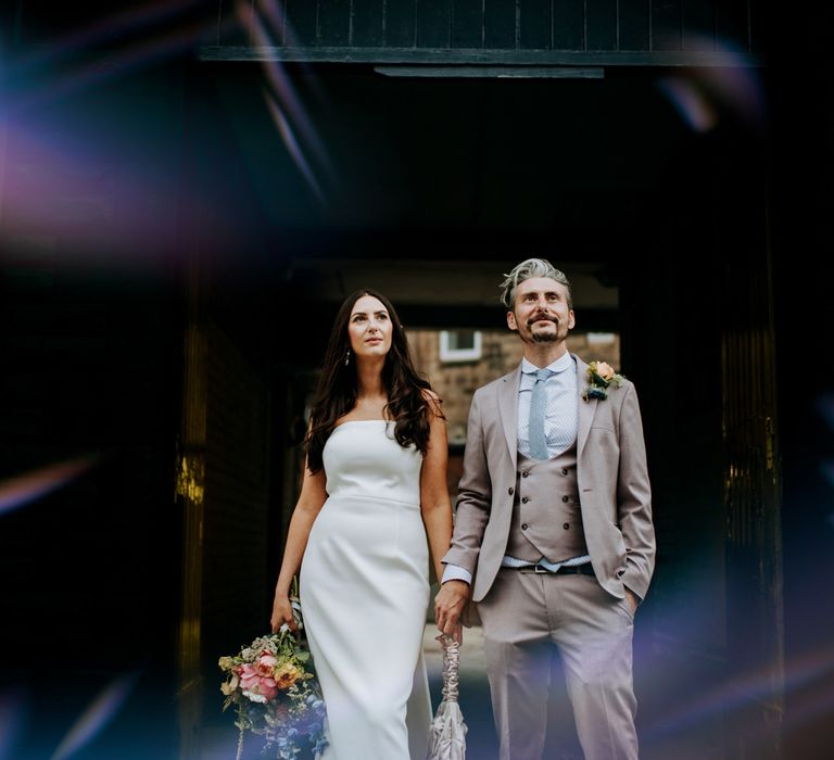 Bride in white strapless Rebecca Vallance Dress holds ivory bridal bag and multicoloured bridal bouquet next to groom in brown Moss Bros suit and blue tie in Harrogate after wedding ceremony