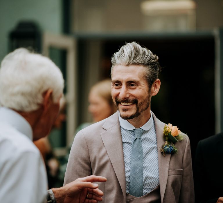 Groom in light brown Moss Bros suit, blue patterned Shelby & Sons shirt, blue tie and multicoloured button hole smiles at wedding guest at Hotel du Vin wedding reception in Harrogate