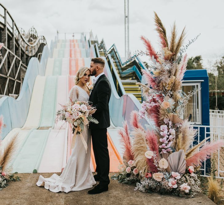 Bride and groom standing next to the Astro slide at Dreamgate Margate with vertical flower arrangements with coloured pampas grass, anthuriums and dried palm leaves 