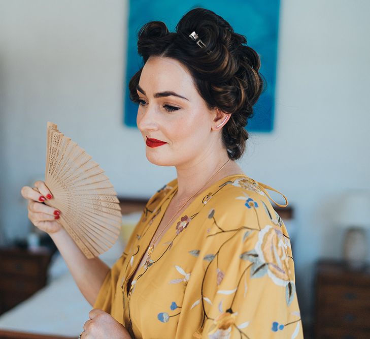 Bride has her hair pinned into curls on her wedding day as she fans her face and wears yellow dressing gown