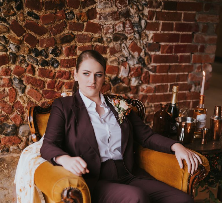 Bride in a plum coloured suit with black high heels sitting in a vintage arm chair for pumpkin spice wedding theme