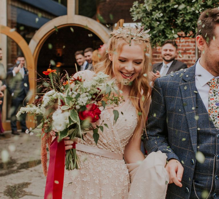 Smiling bride in pearl wedding dress holding red and green bridal bouquet walks holding hands with groom as guests throw confetti after barn wedding ceremony 