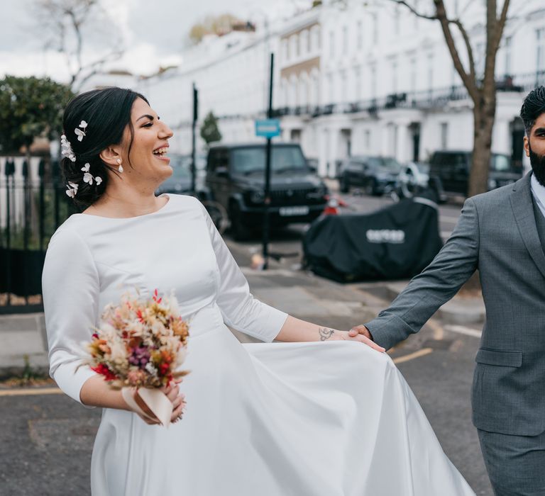 Bride lifts her wedding gown up as she laughs and holds dried floral bouquet whilst walking through the streets of Chelsea