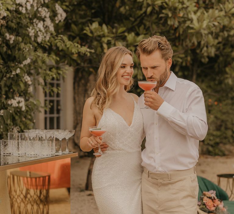 Groom in a white open collar shirt and beige trousers sipping cocktails with his bride in a sparkly Made With Love wedding dress