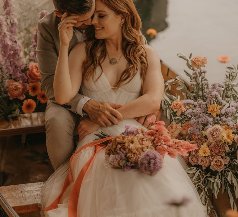 Groom in a beige suit embracing his bride in a Made With Love wedding dress as they sit on a boat at Euridge Manor surrounded by pastel wedding flowers 